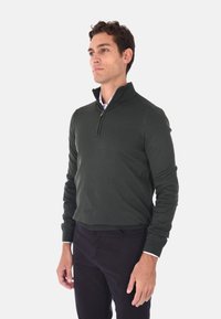 Maglione verde scuro con colletto a zip, maniche lunghe e polsini a costine. Indossato sopra una camicia bianca con colletto e abbinato a pantaloni scuri.