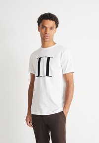 Camiseta de algodón blanca con un audaz diseño en negro de "II" en el frente. Mangas cortas con un cuello redondo clásico, combinada con pantalones marrones.