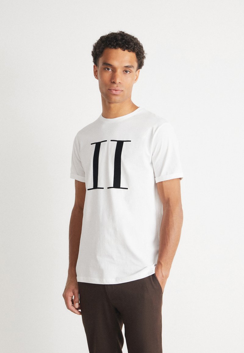 Camiseta de algodón blanca con un audaz diseño en negro de "II" en el frente. Mangas cortas con un cuello redondo clásico, combinada con pantalones marrones.