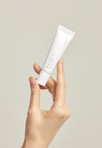 Tubetto di prodotto per labbra bianco tenuto in mano, con texture liscia, tappo piatto e marchio minimalista in grigio.