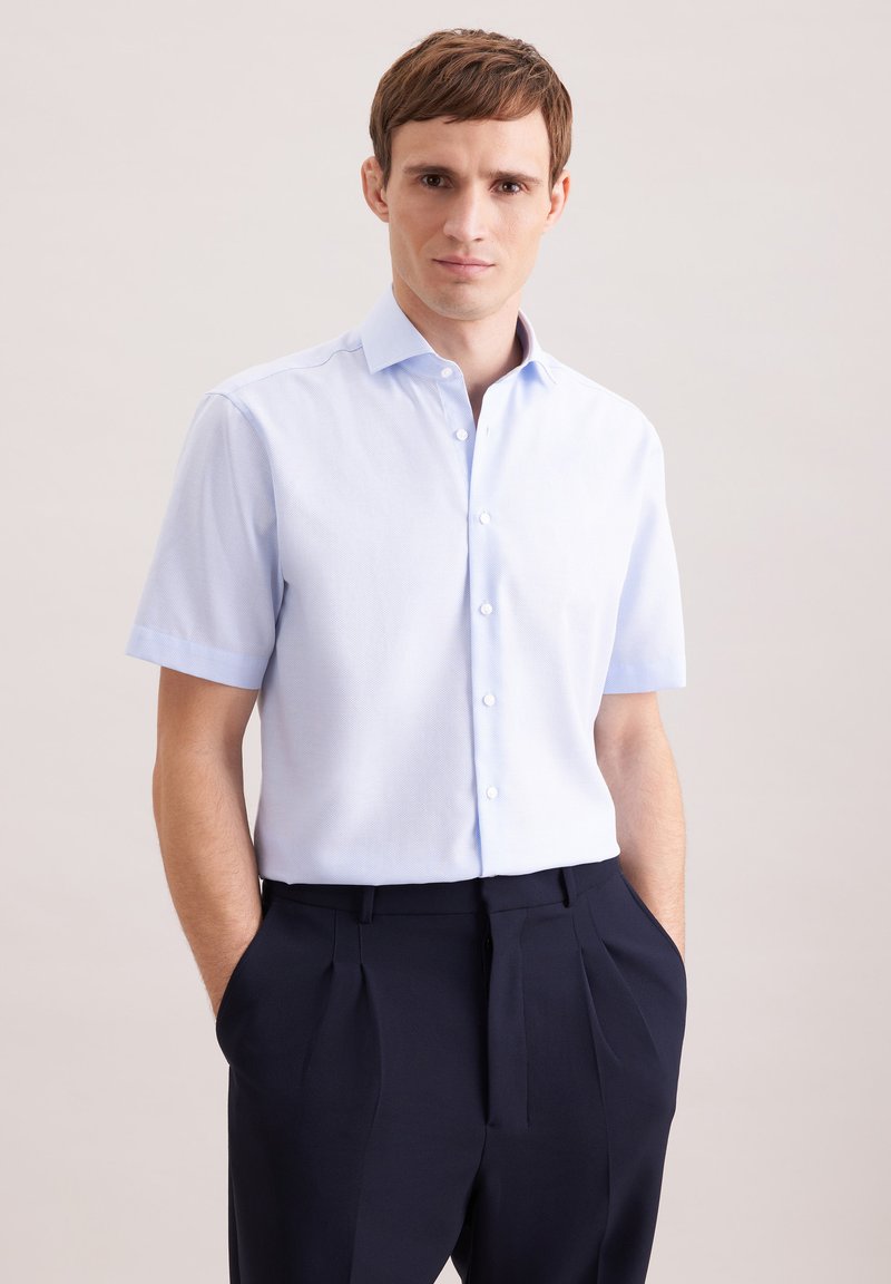 Homme portant une chemise à manches courtes bleu clair, boutonnée et rentrée dans un pantalon noir à plis, les mains dans les poches.
