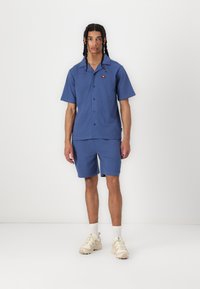 Chemise bleu clair à manches courtes avec des boutons �à l'avant et un petit logo, associée à un short assorti. Portée avec des baskets crème et des chaussettes blanches.