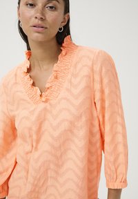 Blouse en pêche avec volants, présentant un motif texturé en zigzag, des manches trois-quarts et un col smocké. Tissu doux et léger.