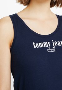 Granatowy bezrękawnik wykonany z bawełny, z białym napisem "tommy jeans" i detalem logo na przodzie, gładka tekstura, okrągły dekolt.