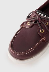 Μπορντό δερμάτινο σκαμπό με λευκή καουτσούκ σόλα, αντίθετα χρυσά μάτια και σκούρες κορδόνες; διαθέτει ραφές κατά μήκος του δακτύλου και των πλαϊνών.