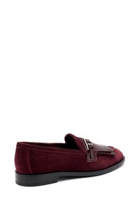 Mocasines de ante burdeos con un pompón de cuero brillante y detalle de hebilla, con una suela baja de goma negra y textura suave.