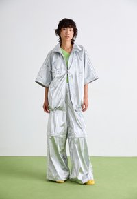 Persoon die op een groene vloer staat en brede zilveren broek draagt met een oversized zilveren jas over een groene top en gele schoenen tegen een witte muur.