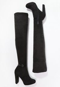 Botas de gamuza negra, por encima de la rodilla, con tacón; una mostrando el lado exterior con hebilla y la otra mostrando el lado interior con cremallera.