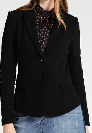 Woman wearing a tailored black blazer over a black blouse with orange polka dots and a tied bow, paired with a blue denim skirt.