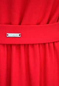 Czerwona sukienka z tkaniny o gładkiej powierzchni, z plisami i srebrnym metalowym znaczkiem przymocowanym do pasa. Prosty i elegancki fason.