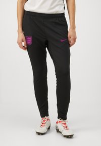 Čierne športové nohavice s úzkym strihom, s fialovým logom Nike a erbom Anglicka; vyrobené z hladkého, ľahkého materiálu.