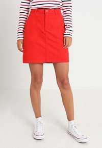 Jupe trapèze en velours côtelé rouge avec fermeture à boutons à l'avant, présentant une texture lisse et une coupe au niveau du genou, assortie à des baskets montantes blanches.