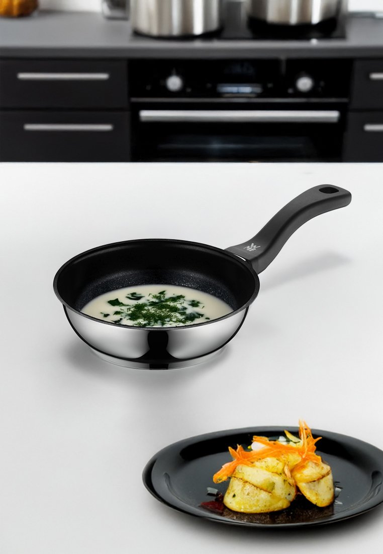 Sartén de acero inoxidable con un interior antiadherente negro, que contiene sopa de crema decorada con hierbas picadas; plato negro con verduras en capas.
