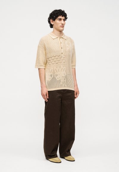 Άνδρας που φοράει μπεζ πλεκτό πόλο με μισά μανίκια και σχέδια, σκούρο φαρδύ παντελόνι και μπεζ μοκασίνια μπροστά σε απλό φόντο.