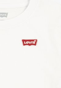 Balti trumpomis rankovėmis medvilninė marškinėliai su raudonu siuvinėtu Levi's logotipu kairėje krūtinės pusėje. Apvalios kaklo formos dizainas.
