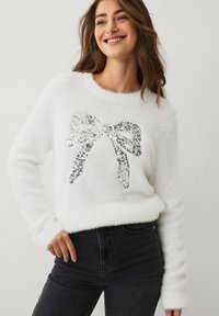 Witte, pluizige trui met een groot zilveren pailletten strikontwerp aan de voorkant. Lange mouwen en een ronde hals. Draagt bij een donkere spijkerbroek.