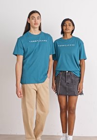 Teal puuvillased T-särgid, millel on "TOMMY JEANS" logo, koos musta teksaskleidiga. Mehemudel kannab beeži pükse; mõlemad mudelid seisavad rahulikult.