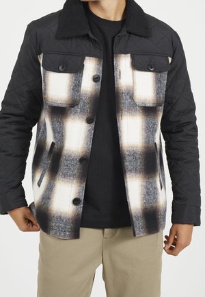 Mann trägt schwarze gesteppte Ärmel und Kragenjacke mit schwarzer, weißer und brauner kariertem Vorderteil über schwarzem Hemd und beigen Hosen.