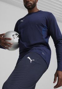 Marinefarbener Langarm-Athletik-Oberteil mit seitlichen Mesh-Details, kombiniert mit passenden Hosen, die ein weißes Logo tragen. Spieler hält einen Fußball.