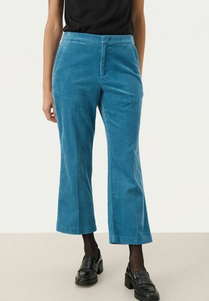 Femme portant un pantalon court en velours côtelé bleu, des collants noirs transparents à pois, des mocassins noirs épais et un haut noir sans manches.