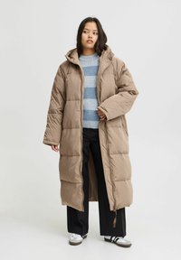 Giacca piumino color marrone chiaro, dotata di cappuccio, maniche lunghe, zip frontale e texture trapuntata. Indossata sopra un maglione a righe blu e pantaloni neri.
