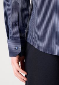 Main reposant près de la hanche, portant une chemise bleu marine à fines rayures avec un poignet boutonné et un pantalon sombre sur fond clair.