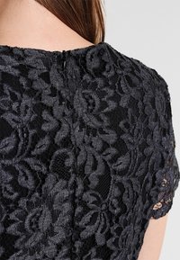 Schwarzes Spitzenoberteil mit aufwendigen Blumenmustern, kurzen Ärmeln und einem Reißverschluss hinten. Die Textur ist zart und semi-transparent.