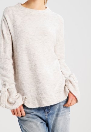 Femme portant un pull en maille beige clair avec des poignets noués et un jean bleu usé, debout devant un fond uni.