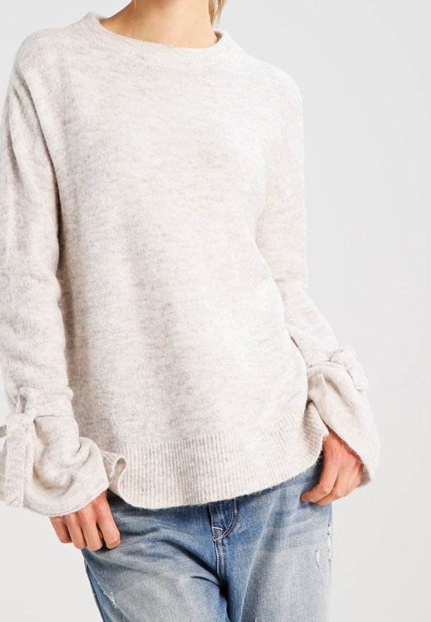 Femme portant un pull en maille beige clair avec des poignets noués et un jean bleu usé, debout devant un fond uni.