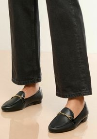 Mocasini negri cu detalii din lanț aurit, purtați cu pantaloni largi închiși la culoare, stând pe o suprafată netedă de culoare deschisă.