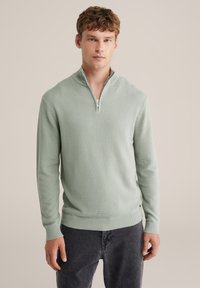 Svijetlozeleni džemper s patentnim zatvaračem i teksturiranim uzorkom pletenja, dugim rukavima i rebrastim manžetnama. Uparen s tamnosivim trapericama.