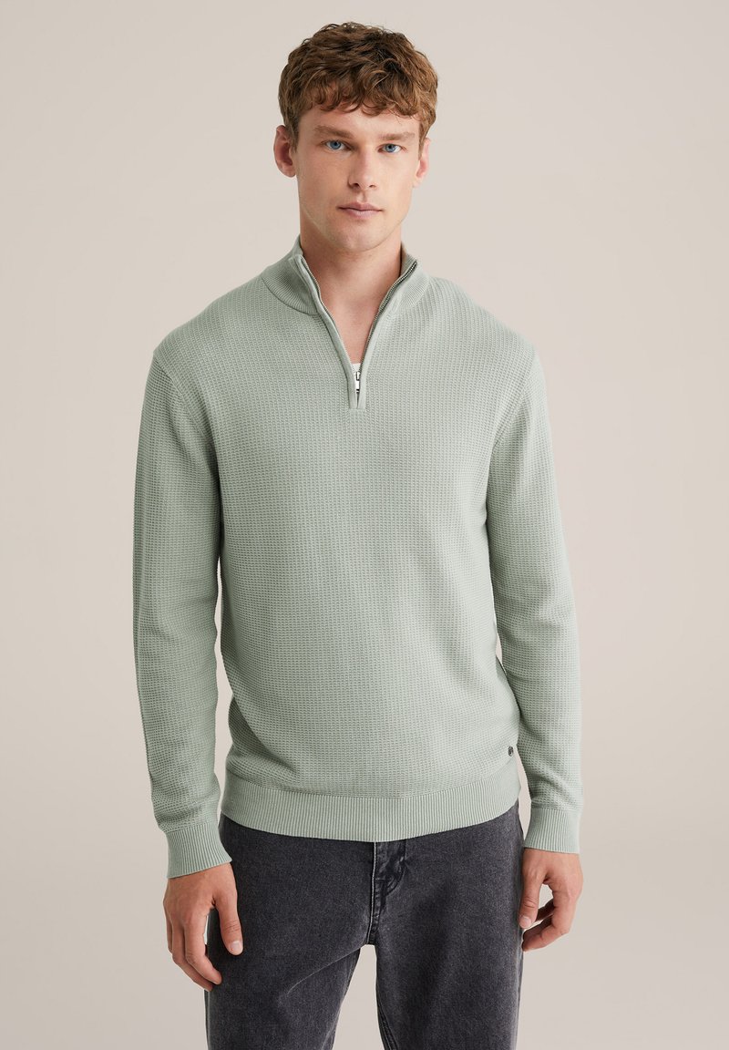 Svijetlozeleni džemper s patentnim zatvaračem i teksturiranim uzorkom pletenja, dugim rukavima i rebrastim manžetnama. Uparen s tamnosivim trapericama.