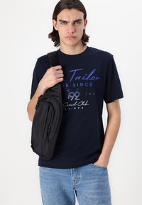 Svart crossbody-veske med flere glidelåslommer, båret over en marineblå t-skjorte med tekst og tall i kontrastfarger.
