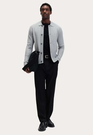 Cardigan gris à col, chemise noire, pantalon noir et chaussures noires. Modèle tenant un sac noir. Texture lisse et boutons visibles.