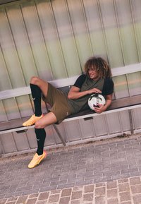 Camiseta deportiva verde y negra con pantalones cortos, calcetas negras y zapatos de fútbol amarillos brillantes; sosteniendo un balón de fútbol en blanco y negro.