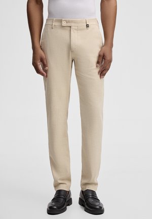 Uomo che indossa pantaloni beige a gamba dritta, camicia bianca infilata e mocassini neri in pelle, in piedi contro uno sfondo semplice.