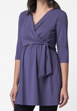 Paarse zwangerschaps top met V-hals, driekwart mouwen, ceintuurdetail in de taille en gladde stof. Gecombineerd met zwarte onderkleding.