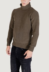 Uomo che indossa un maglione a collo alto a coste di oliva scuro e jeans neri sbiaditi, in piedi contro uno sfondo chiaro e semplice.