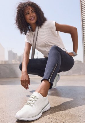 White knit sneakers with light gray accents, rounded toe, and flexible sole. Worn with dark denim jeans featuring zippered cuffs.