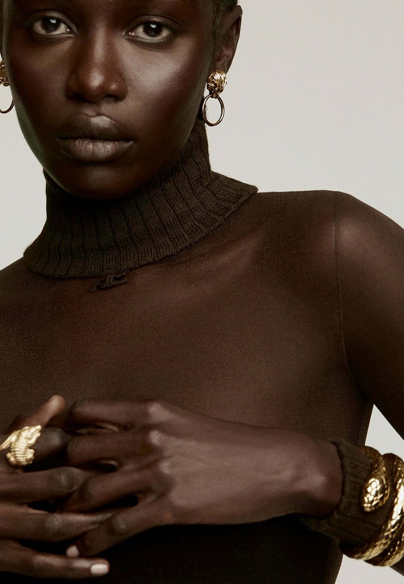 Suéter de cuello alto marrón con un diseño ajustado, acompañado de pendientes y pulseras en tonos dorados. El tono de piel es profundo y rico. Las manos están elegantemente posadas.