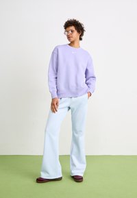 Violets krāsas džemperis ar apaļu izgriezumu un baltu logo, saskaņots ar gaiši zilām izplatītām biksēm. Modelis valkā apaļas brilles un bordo krāsas kurpes.