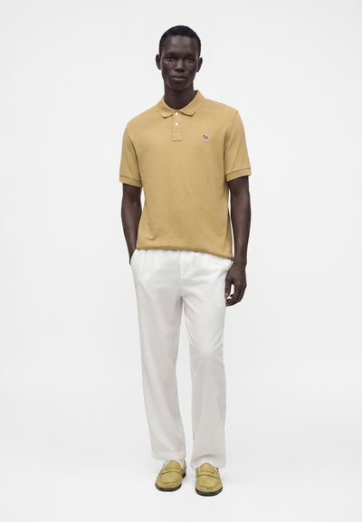 Homme portant un polo jaune moutarde, un pantalon blanc et des mocassins jaune moutarde, debout devant un fond blanc uni.