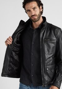 Schwarze Lederjacke mit strukturierten Akzenten, Reißverschluss-Brusttasche, abnehmbarer Fütterung und sichtbaren Nähten, getragen über einem dunklen Hemd mit Knöpfen.
