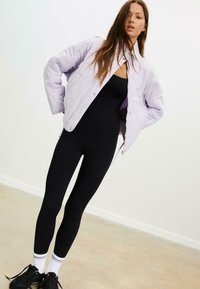 Lichtpaarse gewatteerde bomberjack met een ontspannen pasvorm, gecombineerd met zwarte strak zittende leggings en zwarte sneakers met witte accenten.