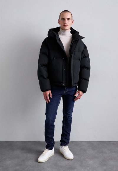Die schwarze gepolsterte Jacke hat eine Kapuze, einen Reißverschluss vorne und zwei Brusttaschen. Sie wird über einem hellen Rollkragenpullover getragen, kombiniert mit blauen Jeans und weißen Sneakers.