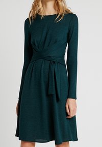 Tmavě modrozelené šaty dlouhého rukávu s přiléhavým korzetem a opaskem v pase. Tkanina má měkkou strukturu a mírně pruží, sukně je zavalená.