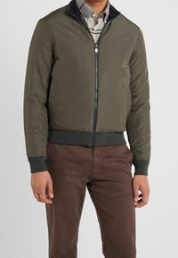 Giacca bomber verde oliva con chiusura a zip, polsini a costine e una texture leggermente lucida. Indossata sopra una camicia a quadri con motivo.