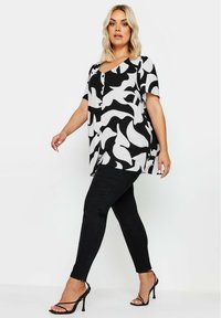 Schwarzes und weißes gemustertes Oberteil mit kurzen Ärmeln, V-Ausschnitt und Knöpfen, kombiniert mit schwarzen schmalen Jeans und schwarzen, geschlossenen High Heels mit Riemen.