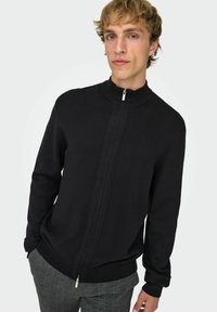 Maglione nero con zip, collo alto, tessuto a maglia, maniche lunghe e polsini a costine. Abbinato a pantaloni grigi a quadri.