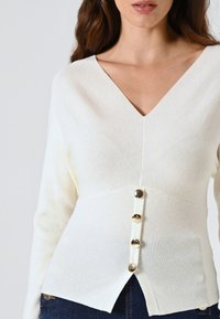 Maglione lavorato a maglia color crema con scollatura a V profonda, vita aderente e quattro bottoni dorati sul davanti, caratterizzato da un design con orlo spaccato.